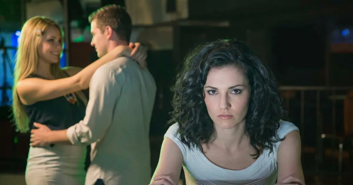 A woman with dark curly hair sits with her arms crossed at a dimly lit bar, looking sad and directly at the camera. In the blurred background, a man intimately embraces a different, blonde woman.